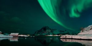 Grüne Polarlichter über verschneiter Küstenlandschaft mit Bergen und Häusern am Wasser